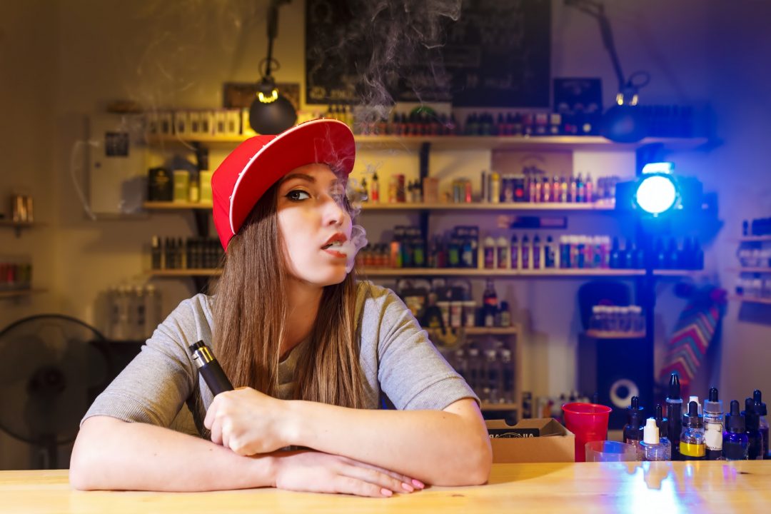 Young pretty woman in red cap smoke an electronic cigarette at the vape shop